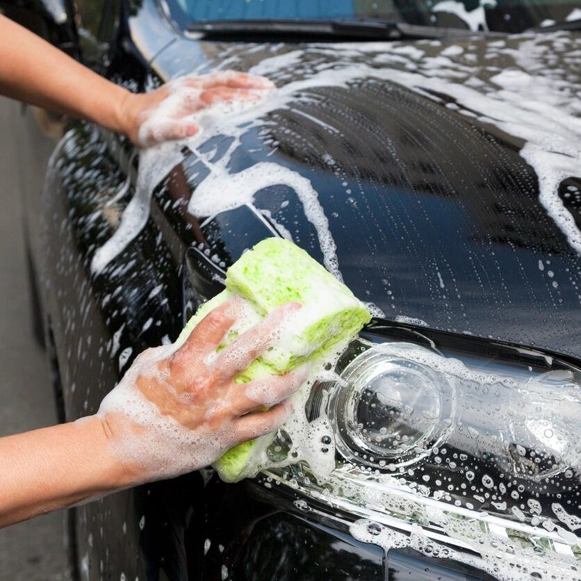 A person washing a black car with soapy water using a green sponge.